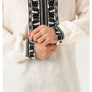May include: Cream-colored linen shirt with intricate black floral embroidery. The shirt features long sleeves with embroidered cuffs and a decorative tassel. A traditional garment with a detailed, symmetrical design.