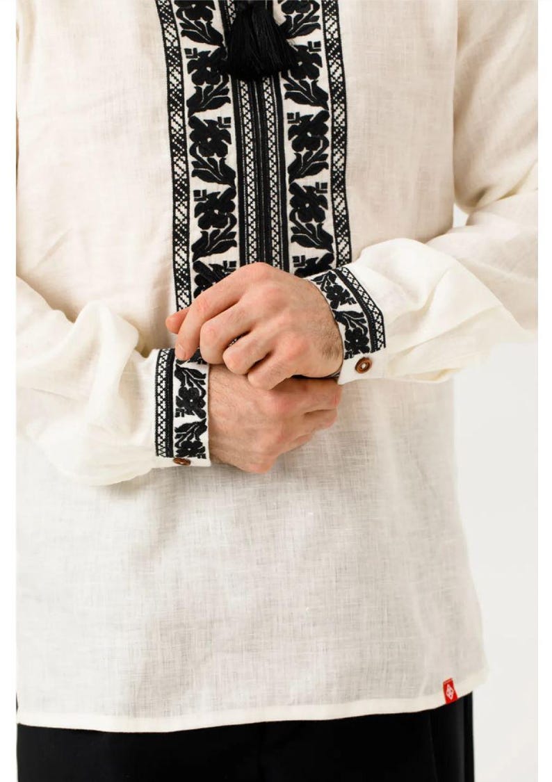 May include: Cream-colored linen shirt with intricate black floral embroidery. The shirt features long sleeves with embroidered cuffs and a decorative tassel. A traditional garment with a detailed, symmetrical design.
