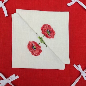 May include: Two red embroidered poppies on a white linen tea towel folded in half. The towel is on a red linen background with white lace trim.