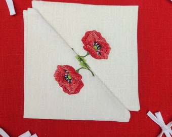 Op de afbeelding: Twee rode geborduurde papavers op een witte linnen theedoek gevouwen in de helft. De theedoek ligt op een rode linnen achtergrond met witte kant.