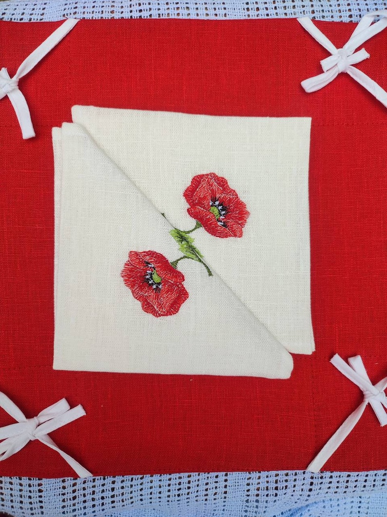 May include: Two red embroidered poppies on a white linen tea towel folded in half. The towel is on a red linen background with white lace trim.