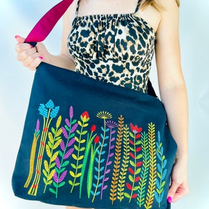 K&ouml;nnte beinhalten: Eine schwarze Canvas-Tragetasche mit einem bunten, gestickten Blumenmuster. Die Tasche hat einen burgunderfarbenen Riemen.