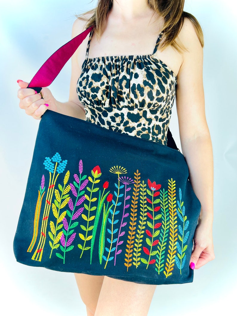 K&ouml;nnte beinhalten: Eine schwarze Canvas-Tragetasche mit einem bunten, gestickten Blumenmuster. Die Tasche hat einen burgunderfarbenen Riemen.