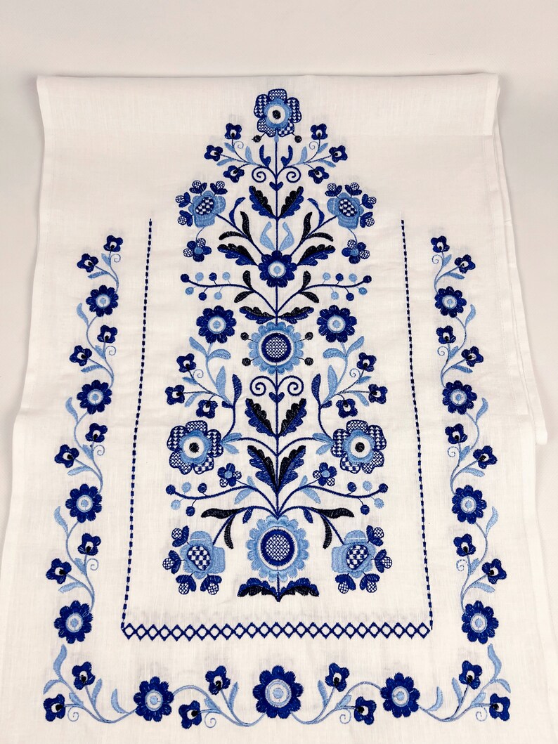 May include: White linen tea towel with blue and black embroidered floral design. The design features a central floral motif with smaller floral motifs along the sides.