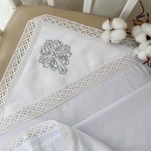 May include: White baby blanket with a gray embroidered cross design and white lace trim.