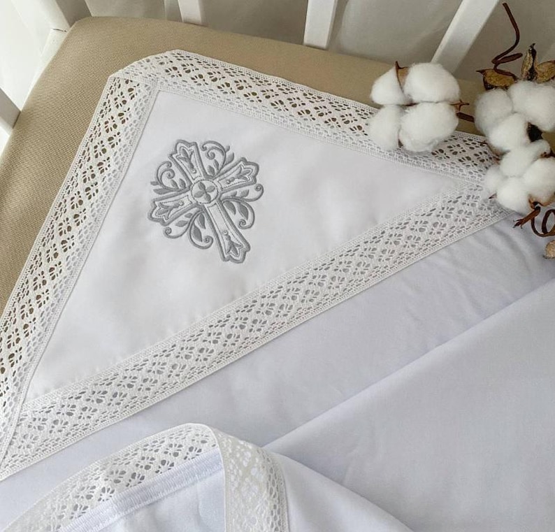 May include: White baby blanket with a gray embroidered cross design and white lace trim.