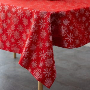 May include: A red tablecloth with a repeating pattern of white snowflakes. The tablecloth is draped over a table, with the corner of the cloth hanging down. The fabric has a slight sheen, and the snowflakes are detailed.
