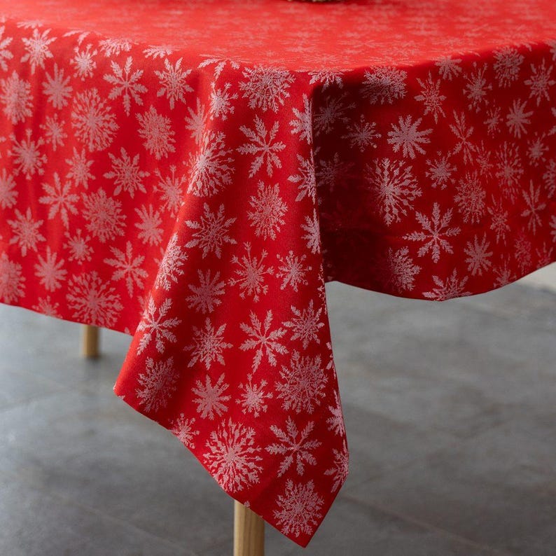May include: A red tablecloth with a repeating pattern of white snowflakes. The tablecloth is draped over a table, with the corner of the cloth hanging down. The fabric has a slight sheen, and the snowflakes are detailed.