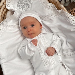 Op de afbeelding: Een witte baby-outfit met een kanten rand en een witte muts. De outfit ligt op een witte deken met een kanten rand en een zilveren kruisborduursel.