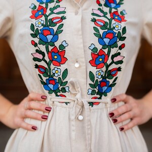 May include: A beige linen dress with vibrant floral embroidery in red, blue, and green. The embroidery adorns the bodice and sleeves, with a tie waist and button-down front. The dress has long sleeves and a collared neckline.