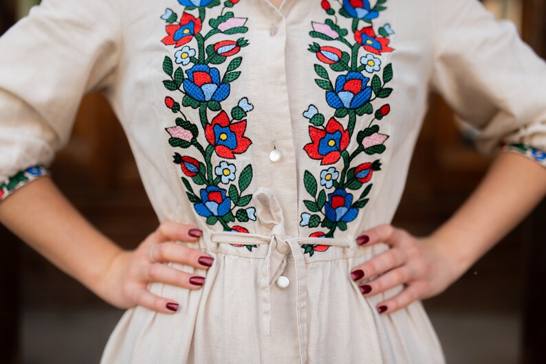 May include: A beige linen dress with vibrant floral embroidery in red, blue, and green. The embroidery adorns the bodice and sleeves, with a tie waist and button-down front. The dress has long sleeves and a collared neckline.