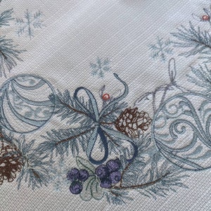 K&ouml;nnte beinhalten: Nahaufnahme eines hellen Stoffes mit einem wiederholenden, weihnachtlichen, gestickten Rand. Der Rand zeigt blau-gr&uuml;ne Tannenzweige, Ornamente, Tannenzapfen und violette Beeren. Der Stoff hat ein subtiles strukturiertes Muster.