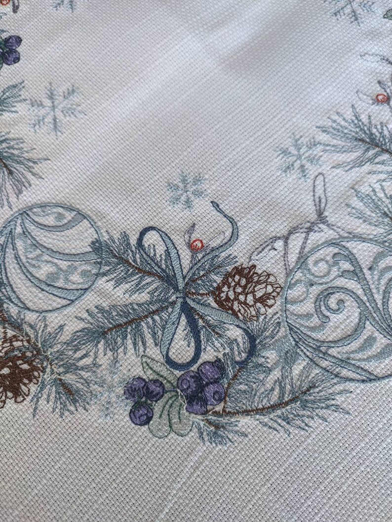 K&ouml;nnte beinhalten: Nahaufnahme eines hellen Stoffes mit einem wiederholenden, weihnachtlichen, gestickten Rand. Der Rand zeigt blau-gr&uuml;ne Tannenzweige, Ornamente, Tannenzapfen und violette Beeren. Der Stoff hat ein subtiles strukturiertes Muster.