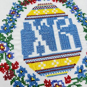 May include: A white fabric with a blue and yellow embroidered egg surrounded by a wreath of red, blue, green, and yellow flowers.