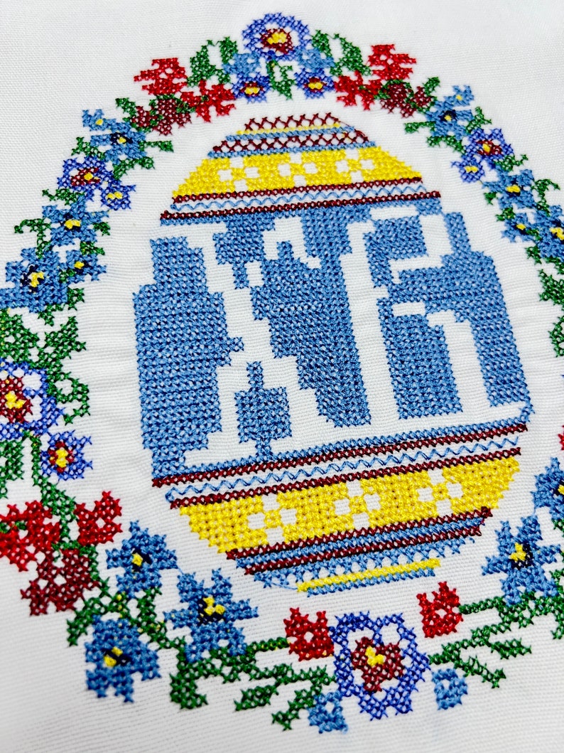 May include: A white fabric with a blue and yellow embroidered egg surrounded by a wreath of red, blue, green, and yellow flowers.