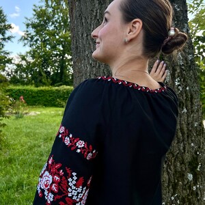 K&ouml;nnte beinhalten: Eine Frau tr&auml;gt eine schwarze bestickte Bluse mit roten und wei&szlig;en Blumendesigns. Die Bluse hat einen runden Ausschnitt und lange &Auml;rmel. Die Frau steht neben einem Baum.