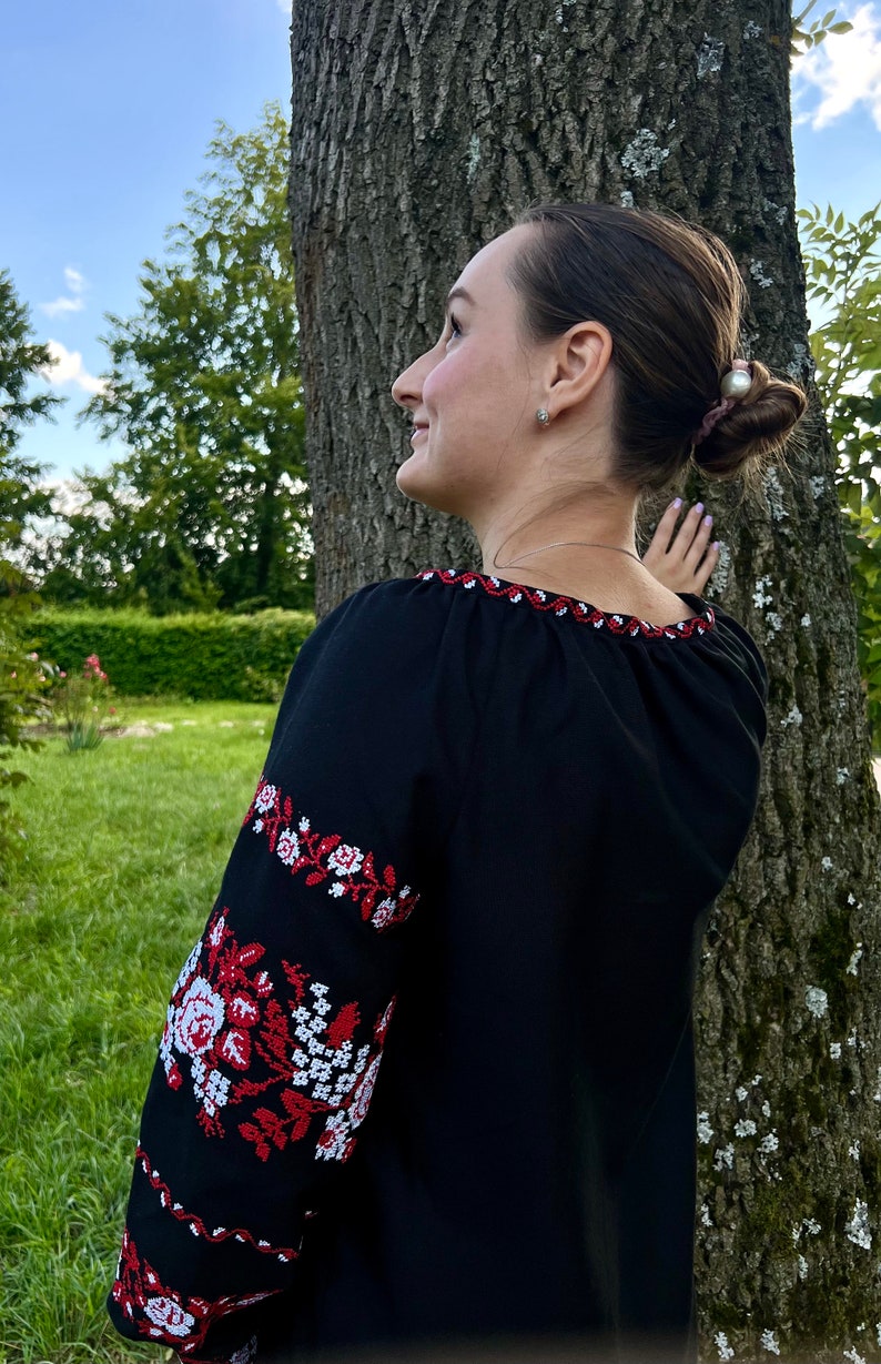 K&ouml;nnte beinhalten: Eine Frau tr&auml;gt eine schwarze bestickte Bluse mit roten und wei&szlig;en Blumendesigns. Die Bluse hat einen runden Ausschnitt und lange &Auml;rmel. Die Frau steht neben einem Baum.