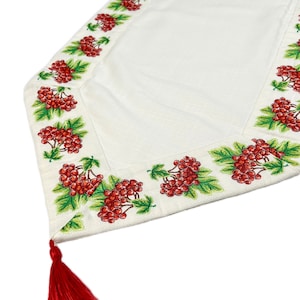 May include: White table runner with a detailed border featuring red berries and green leaves. A red tassel hangs from the corner. The runner is made of a textured fabric, suitable for home decor.