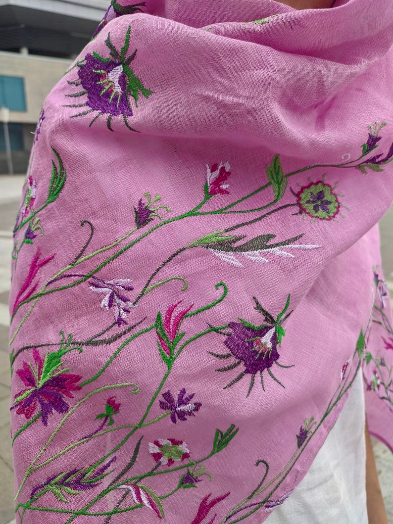 Puede incluir: Una bufanda de lino rosa con dise&ntilde;os florales bordados en tonos de morado, verde y blanco.
