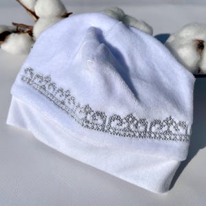 K&ouml;nnte beinhalten: Ein wei&szlig;er Baby-Beanie mit silbernem Stickdesign am Rand. Der Hut ist aus einem weichen, pl&uuml;schigen Material gefertigt und am unteren Ende umgeschlagen. Baumwollzweige befinden sich im Hintergrund.