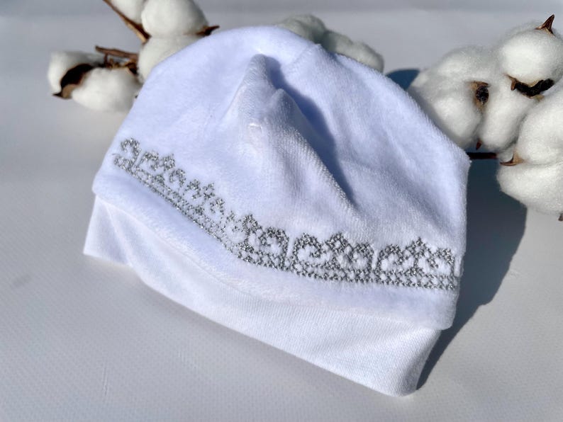 K&ouml;nnte beinhalten: Ein wei&szlig;er Baby-Beanie mit silbernem Stickdesign am Rand. Der Hut ist aus einem weichen, pl&uuml;schigen Material gefertigt und am unteren Ende umgeschlagen. Baumwollzweige befinden sich im Hintergrund.