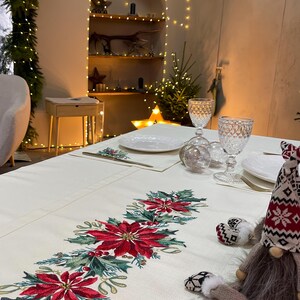 May include: A white tablecloth with a red and green embroidered poinsettia design. A small gnome figurine with a grey fur beard and red and white knit hat sits on the tablecloth.