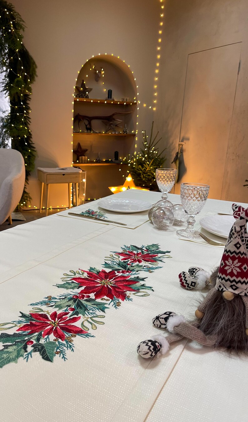 May include: A white tablecloth with a red and green embroidered poinsettia design. A small gnome figurine with a grey fur beard and red and white knit hat sits on the tablecloth.