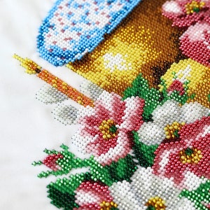 K&ouml;nnte beinhalten: Nahaufnahme eines Perlenblumendesigns mit einem blau-wei&szlig;en Objekt, einer Kerze und einem braun-gelben Objekt. Die Blumen sind rot, wei&szlig; und rosa mit gr&uuml;nen Bl&auml;ttern. Der Hintergrund ist wei&szlig;.