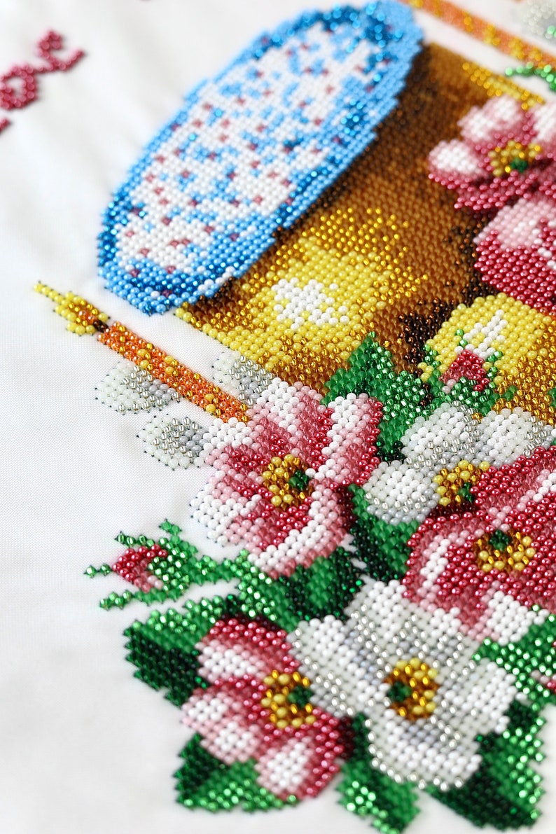 K&ouml;nnte beinhalten: Nahaufnahme eines Perlenblumendesigns mit einem blau-wei&szlig;en Objekt, einer Kerze und einem braun-gelben Objekt. Die Blumen sind rot, wei&szlig; und rosa mit gr&uuml;nen Bl&auml;ttern. Der Hintergrund ist wei&szlig;.