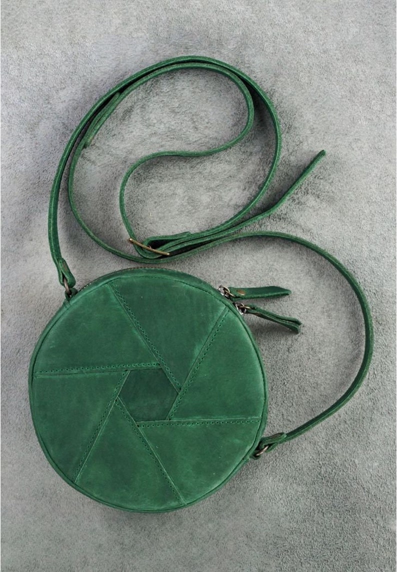 Op de afbeelding: Een ronde, groene leren schoudertas met een geometrisch patroon. De tas heeft een lange verstelbare schouderband en een ritssluiting.