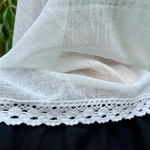 May include: Close-up of a white, sheer fabric with a delicate lace trim. The fabric is layered, revealing a glimpse of a black garment beneath. The lace trim features a scalloped edge, adding a touch of elegance.