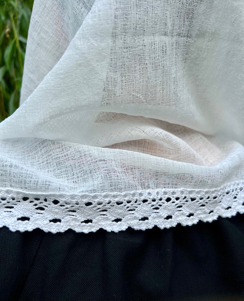 May include: Close-up of a white, sheer fabric with a delicate lace trim. The fabric is layered, revealing a glimpse of a black garment beneath. The lace trim features a scalloped edge, adding a touch of elegance.