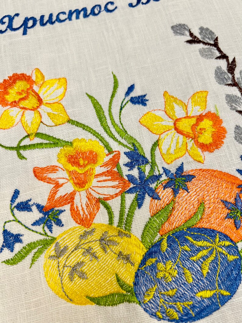 Puede incluir: Dise&ntilde;o de Pascua bordado con flores amarillas, naranjas y azules, hojas verdes y tres huevos de Pascua coloridos. El texto "Христос" est&aacute; bordado en hilo azul en la parte superior del dise&ntilde;o.