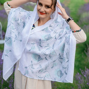 Puede incluir: Un chal de lino blanco con un estampado floral en tonos azules y grises. El chal est&aacute; drapeado sobre la cabeza y los hombros. La persona lleva un vestido beige y un reloj con una correa negra.