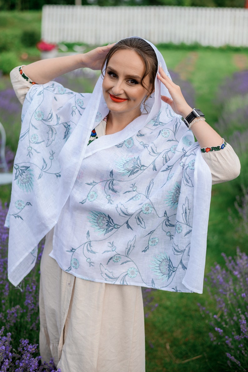 Puede incluir: Un chal de lino blanco con un estampado floral en tonos azules y grises. El chal est&aacute; drapeado sobre la cabeza y los hombros. La persona lleva un vestido beige y un reloj con una correa negra.
