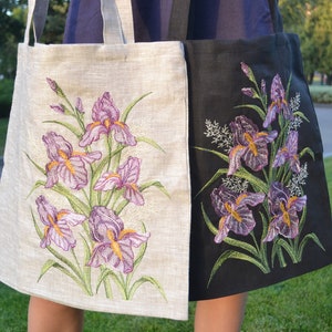 Op de afbeelding: Twee tote bags met geborduurde irisbloemen. E&eacute;n tas is van wit linnen met paarse en groene bloemen. De andere tas is van zwart linnen met paarse en groene bloemen.