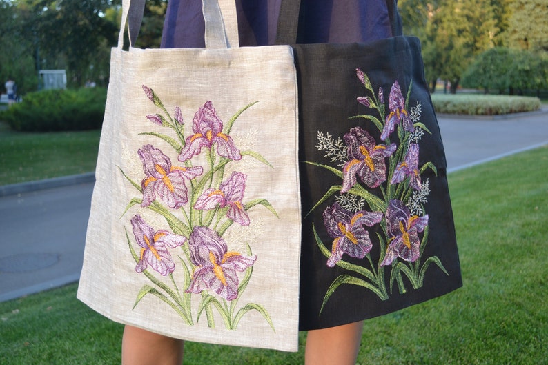 Op de afbeelding: Twee tote bags met geborduurde irisbloemen. E&eacute;n tas is van wit linnen met paarse en groene bloemen. De andere tas is van zwart linnen met paarse en groene bloemen.