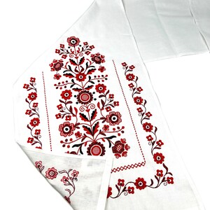 K&ouml;nnte beinhalten: Wei&szlig;er Tischl&auml;ufer aus Leinen mit roten und schwarzen gestickten Blumendesigns. Das Design zeigt ein zentrales Blumenmotiv mit kleineren Blumenbord&uuml;ren.