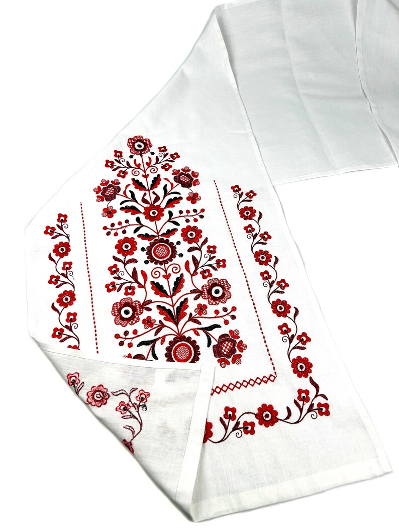 K&ouml;nnte beinhalten: Wei&szlig;er Tischl&auml;ufer aus Leinen mit roten und schwarzen gestickten Blumendesigns. Das Design zeigt ein zentrales Blumenmotiv mit kleineren Blumenbord&uuml;ren.