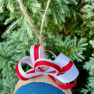 K&ouml;nnte beinhalten: Ein blauer und wei&szlig;er Weihnachtsbaumschmuck mit einer Jutebasis und einer roten und wei&szlig;en Schleife. Der Schmuck ist mit einem Design aus Stechpalme und Beeren verziert.