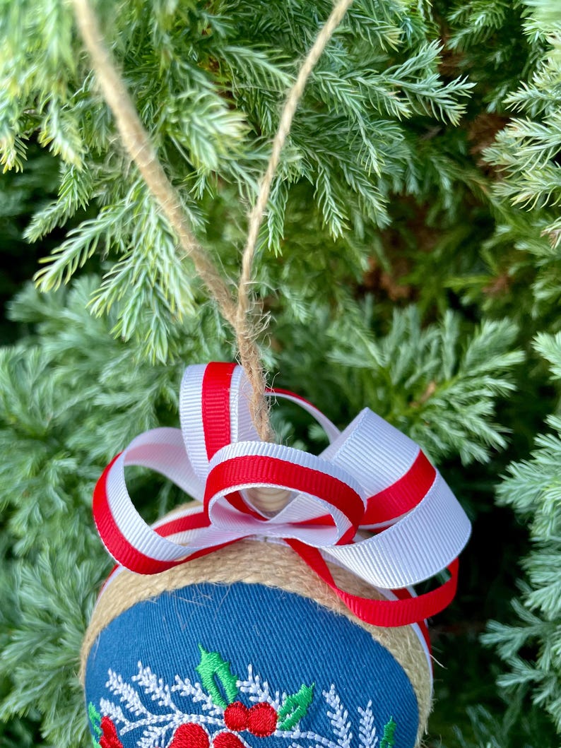 K&ouml;nnte beinhalten: Ein blauer und wei&szlig;er Weihnachtsbaumschmuck mit einer Jutebasis und einer roten und wei&szlig;en Schleife. Der Schmuck ist mit einem Design aus Stechpalme und Beeren verziert.