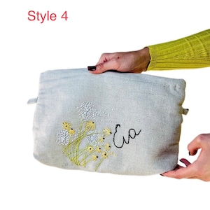 May include: A light gray canvas pouch with a floral embroidery design and the name "Eva" embroidered in black thread.