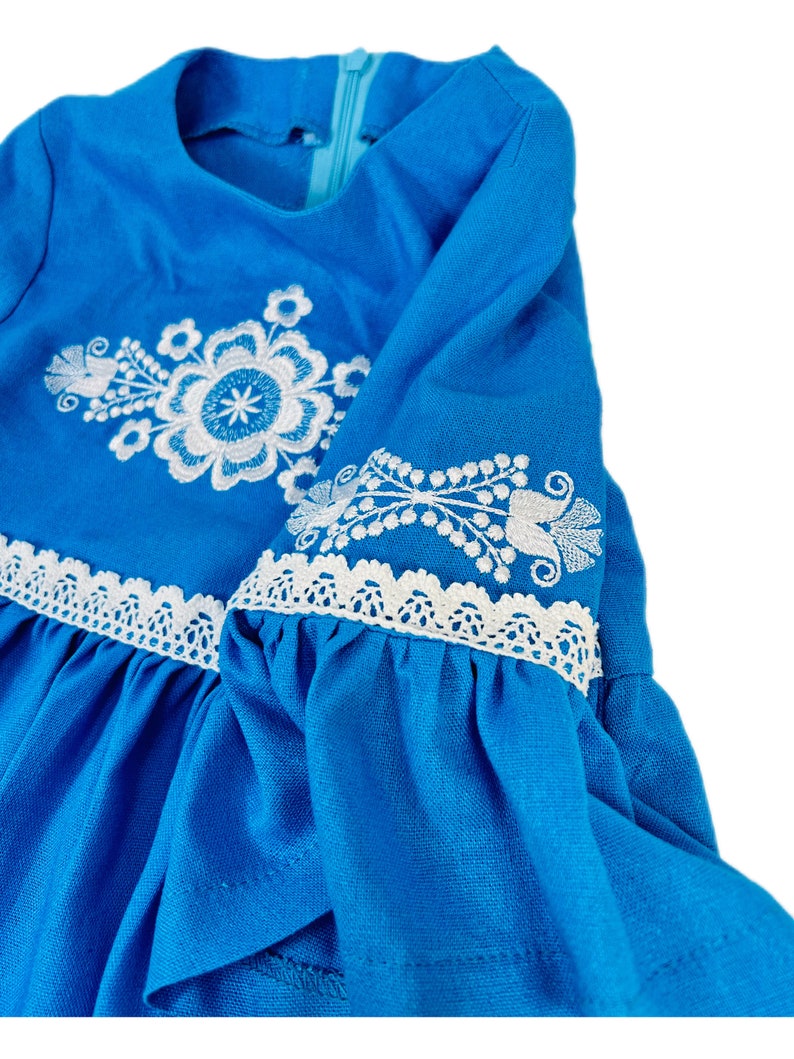 K&ouml;nnte beinhalten: Ein blaues Kleid mit wei&szlig;en gestickten Blumenmustern und wei&szlig;en Spitzenbesatz an den &Auml;rmeln.