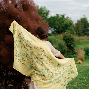 May include: A woman wearing a light yellow shawl with a floral pattern in shades of blue and green. The shawl is draped over her shoulders and she is holding it in her hands. The background is a blurry image of a garden.