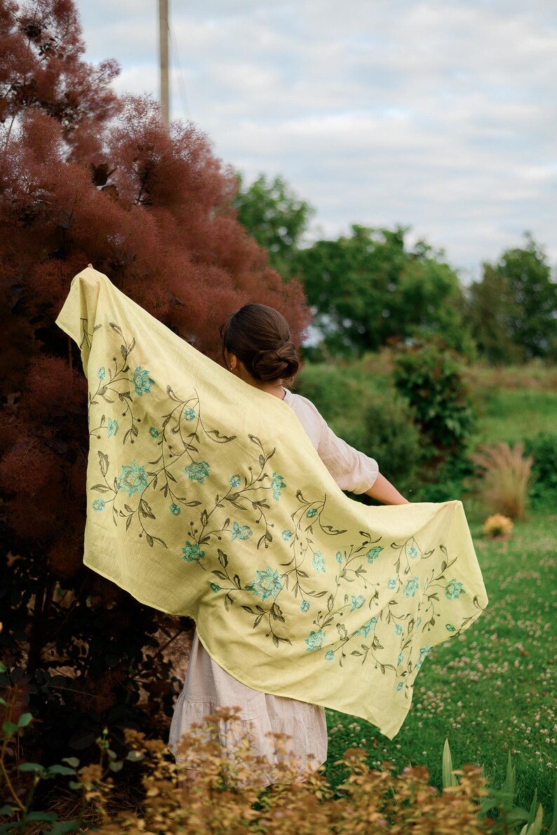 May include: A woman wearing a light yellow shawl with a floral pattern in shades of blue and green. The shawl is draped over her shoulders and she is holding it in her hands. The background is a blurry image of a garden.