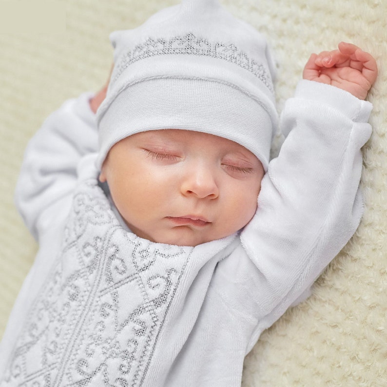 K&ouml;nnte beinhalten: Ein schlafendes Baby in einem wei&szlig;en Outfit und Hut mit silbernen Stickereien. Die Arme des Babys sind erhoben, und die Augen sind geschlossen. Das Outfit scheint aus einem weichen, pl&uuml;schigen Material zu sein. Der Hintergrund ist eine helle, strukturierte Oberfl&auml;che.
