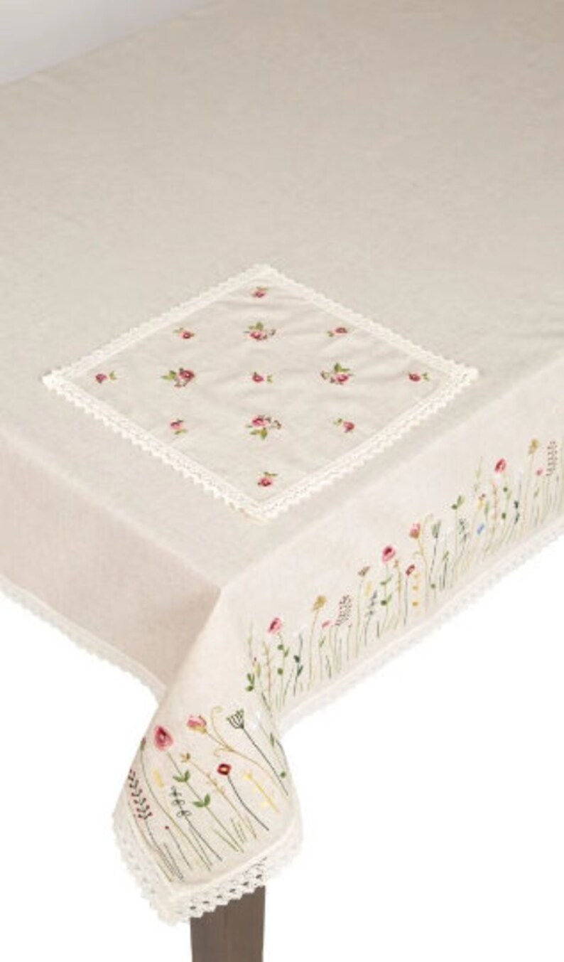May include: Beige linen tablecloth with embroidered floral details. Features a square embroidered centerpiece with pink flowers and a delicate lace trim. The edges are adorned with embroidered wildflowers.