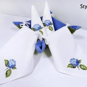 May include: Four white linen napkins with blue hydrangea embroidery. The napkins are folded into cones and placed in a blue basket. The text "Style 3" is visible in the top right corner of the image.
