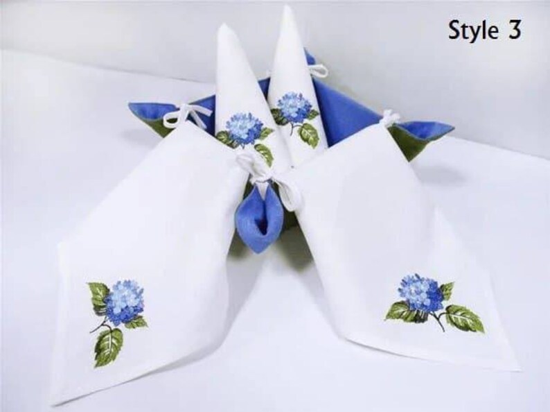May include: Four white linen napkins with blue hydrangea embroidery. The napkins are folded into cones and placed in a blue basket. The text "Style 3" is visible in the top right corner of the image.