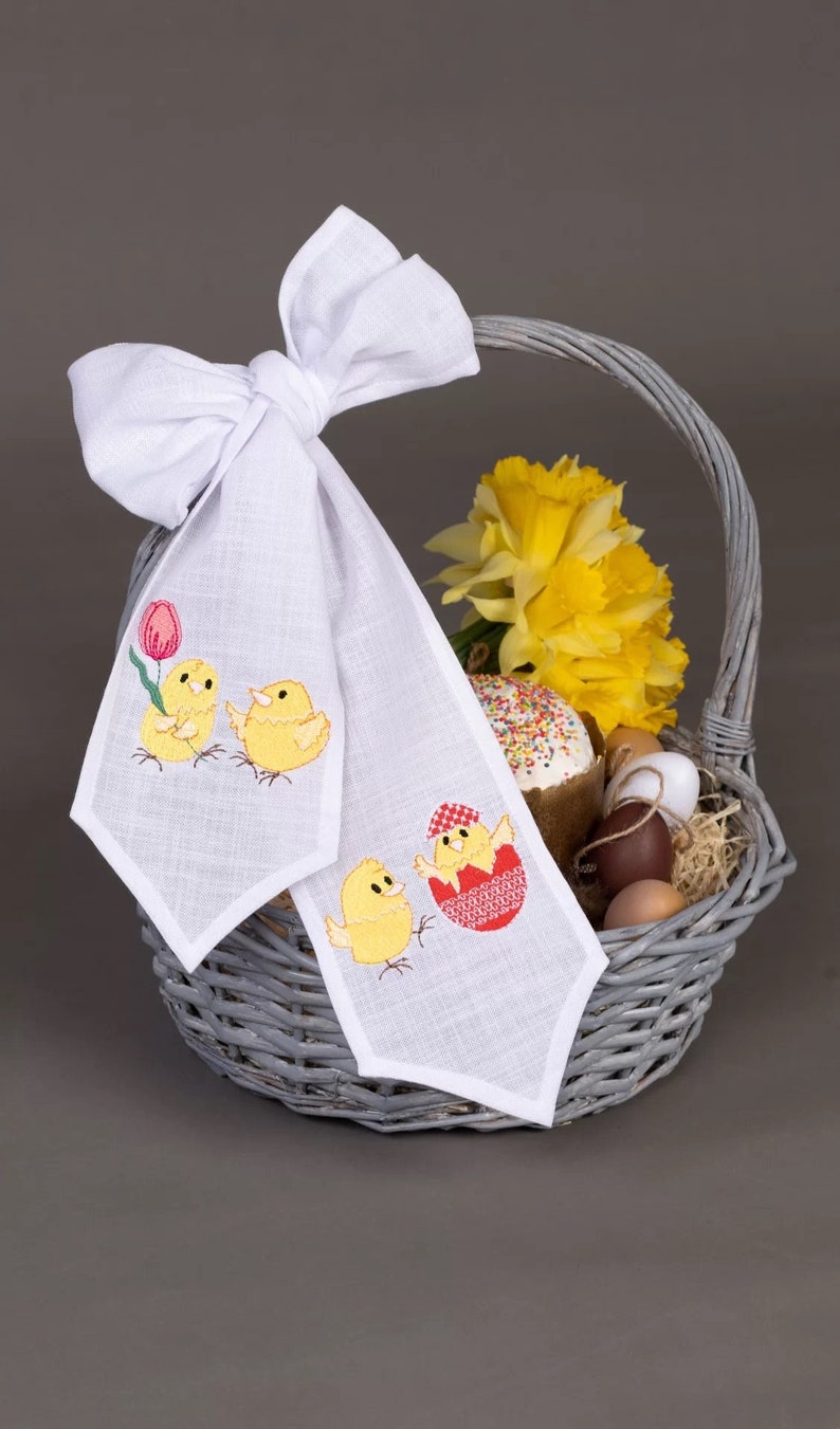 May include: A white linen Easter basket liner with embroidered chicks and a tulip. The liner is tied with a white bow and is filled with a decorated Easter cake, yellow daffodils, and brown eggs.